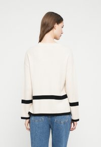 Kobieta z długimi brązowymi włosami, ubrana w kremowy sweter w czarne poziome paski i niebieskie dżinsy, stoi tyłem na jednolitym tle.