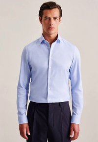 Camicia azzurro chiaro con colletto a punta, maniche lunghe e texture liscia, indossata con pantaloni scuri su misura.