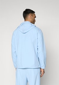 Chaqueta con capucha de color azul claro, hecha de tela ligera. Cuenta con puños elásticos y un corte relajado, con un diseño sencillo y sin patrones visibles.