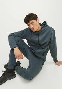 Mørkeblå træningsdragt lavet af blødt stof, med en lynlåset hoodie og elastiske manchetter, parret med sorte træningssko. Glat tekstur, afslappet pasform.
