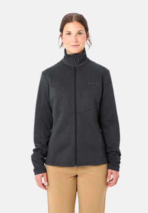 Vrouw met bruin haar, gekleed in een donkergrijze, gestructureerde jas met volledige ritssluiting en beige broek, staand tegen een effen lichte achtergrond.