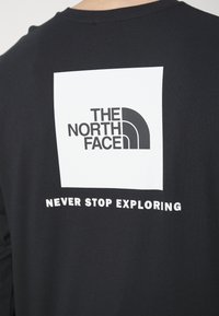 Black long-sleeve shirt featuring a white rectangular graphic with "THE NORTH FACE" logo and "NEVER STOP EXPLORING" text below.