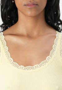 Débardeur jaune à côtes avec un col en dentelle à volants. Texture lisse avec une délicate bordure en dentelle, mettant en avant un design subtil et élégant.