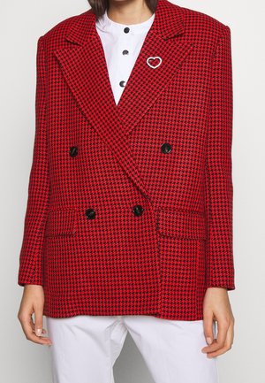 Kobieta w czerwono-czarnej marynarce dwurzędowej w kratę psy z czarnymi guzikami, białej koszuli z czarnymi guzikami oraz białych spodniach.