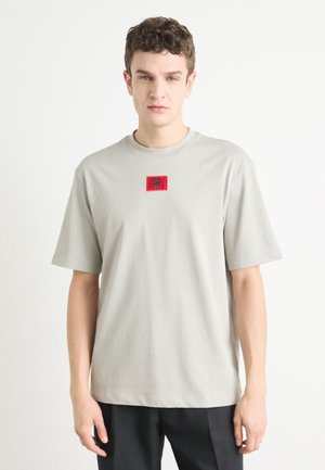 T-shirt en coton gris avec un col rond, des manches courtes et un patch logo carré rouge sur la poitrine. Texture lisse, coupe décontractée.