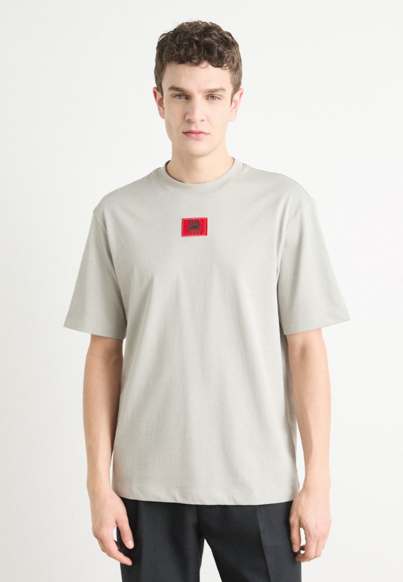 T-shirt en coton gris avec un col rond, des manches courtes et un patch logo carré rouge sur la poitrine. Texture lisse, coupe décontractée.