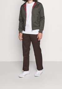 Giacca bomber verde con rivestimento a quadri, T-shirt bianca, pantaloni marroni e sneaker bianchi. Presenta tasche e polsini a costine.