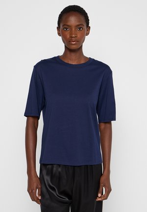 Kobieta ubrana w gładki granatowy t-shirt z krótkim rękawem i czarne satynowe spodnie, stojąca na jednolitym jasnoszarym tle.