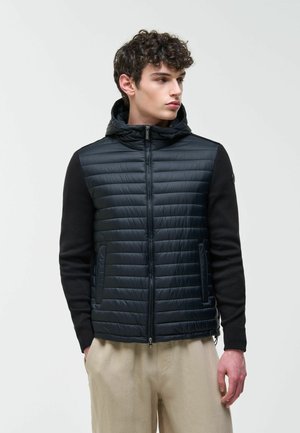 Mladý muž s kučeravými vlasmi, oblečený v čiernej prešívanej bunde s kapucňou a čiernymi rukávmi, s béžovými nohavicami, hľadiaci doprava.