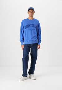Modra trenirka z logotipom "TOMMY JEAN New York", kombinirana s temnimi jeans hlačami in belimi supergami. Trenirka ima rebraste manšete in spodnji rob.