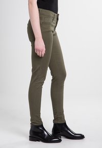 Olive krāsas šaurās džinsi, izgatavoti no elastīgas kokvilnas, ar vidēju jostas augstumu, priekšējām kabatām un gludu virsmu, apvienoti ar melnajām potīšu kurpēm.