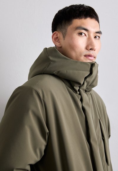 Giacca impermeabile verde oliva con colletto alto, cappuccio grande e design minimalista. Presenta una chiusura con zip e dettagli con tasche laterali.