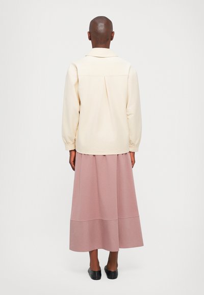 Κρεμ μπλούζα με ελαστικούς μανσέτες, συνδυασμένη με ροζ μακριά φούστα. Το σύνολο έχει χαλαρή εφαρμογή και λεπτομέρεια πτυχής στην πλάτη.