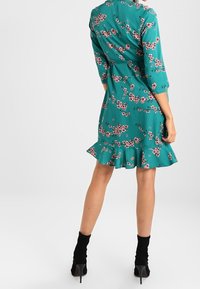 Türkises Kleid mit Dreiviertelärmeln, floralem Muster, schwingendem Saum und figurbetontem Bund. Getragen mit schwarzen Knöchelstiefeln.