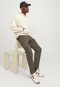 Hanorac bej cu șnur, pantaloni cargo olive cu buzunare laterale și adidași crem cu accente bordeux, așezat pe un scaun deschis la culoare.