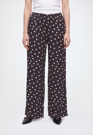 Modèle portant un pantalon noir à jambes larges à pois blancs, un haut blanc et des chaussures noires pointues.