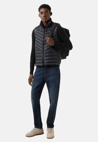 Schwarze gesteppte Steppweste mit horizontalen Nähten, kombiniert mit einem schwarzen Langarmshirt, dunkler Jeans und beigen Turnschuhen. Grauer Rucksack über der Schulter.