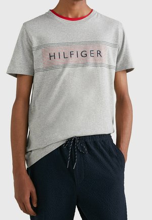 Person iført en lysegrå T-shirt med teksten "HILFIGER" og sorte snor mørkeblå bukser, hænder afslappet ved siden.