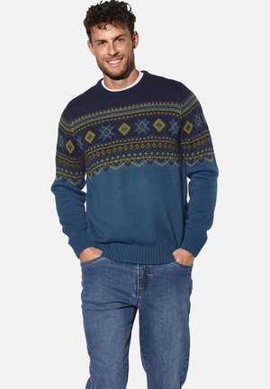 Knit sweater featuring multicolored geometric patterns in navy, green, and blue. Ribbed cuffs and hem with a round neck. Worn with denim jeans.