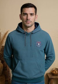 Blauwe gestreepte hoodie met een voorzak, een capuchon met trekkoord en een logo-embleem. Het materiaal lijkt zacht, met ribgebreide manchetten en zoom.