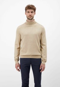 Beige Rollkragenspullover mit gerippter Textur und taillierter Passform, getragen von einem Model vor einem schlichten Hintergrund, kombiniert mit dunkelblauen Hosen.