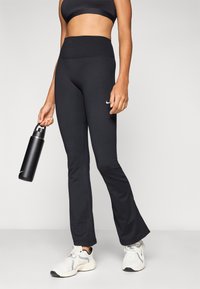 Čierne legíny s rozšírenými nohavicami a vysokým pásom, vyrobené z elastickej látky. Dopĺňané čiernou fľašou na vodu a bielou športovou obuvou.