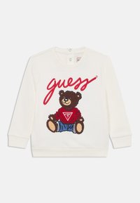 Sweatshirt branco com gola redonda, apresentando um urso castanho bordado com um suéter vermelho e jeans azuis, com a palavra "guess" em letra cursiva vermelha.