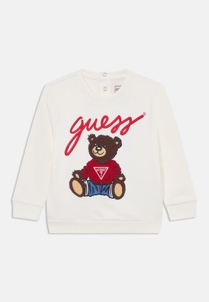 White sweatshirt with round neck, featuring an embroidered brown bear in a red sweater and blue jeans, with the word "guess" in red script.