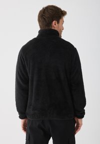 Melna flīsa veste ar augstu apkakli, teksturētu audumu un garām piedurknēm. Tai ir brīva piegriezuma silhouette un tā nosegs muguru.