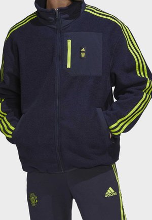 Mann trägt eine marineblaue und neongelbe Adidas Manchester United Fleecejacke und passende Hose, die Hände in den Jackentaschen.