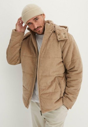 Giacca in corduroy beige con un design voluminoso, chiusura con zip, due tasche laterali e cappuccio staccabile. Indossata con un berretto lavorato beige.