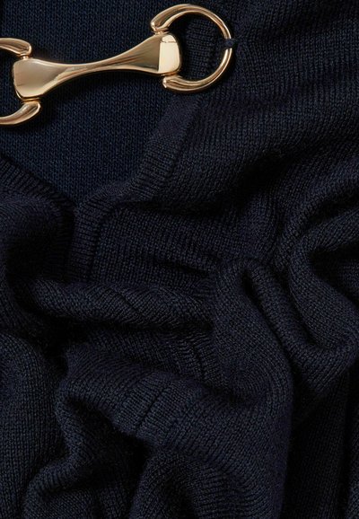 Tessuto a maglia blu navy con una texture morbida, caratterizzato da un accento in hardware dorato. Il tessuto mostra drappeggi e bordi a coste, evidenziando la sua elasticità.