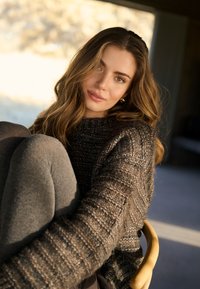 Fiatal nő hosszú göndör hajjal, vastag barna kötött pulóverben és szürke leggingsben, görnyedten ül egy fa széken, beltéren, lágy fény közelében.