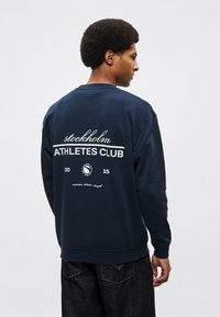 Tmavomodrá mikina s okrúhlym výstrihom, na ktorej je biely nápis "stockholm ATHLETES CLUB" a na zadnej strane dizajn loga. Hladká textúra.
