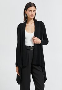 Cárdigan negro con diseño de frente abierto, textura acanalada y mangas largas. Combinado con una blusa blanca ajustada y pantalones negros de cintura alta.
