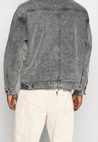 Giacca in denim grigio sbiadito con vestibilità rilassata, cuciture in vista e bottoni a pressione. Indossata con pantaloni in velluto a coste di colore chiaro.