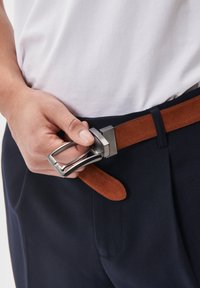 Brun mockaskärp med en silverfärgad rektangulär spänne, bärs över navybyxor. Handen justerar spännet, vilket visar en närbild av materialen.