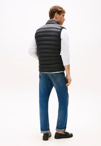 Jovem em pé, vestindo um colete acolchoado preto sobre uma camisa branca de mangas compridas, jeans azuis arregaçados e mocassins pretos, contra um fundo branco.