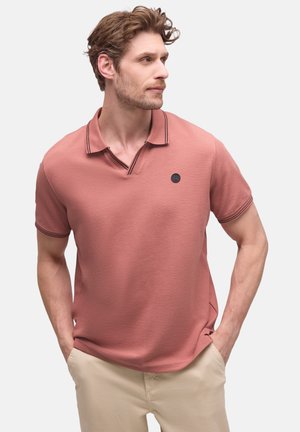 Mężczyzna z jasnobrązowymi włosami, ubrany w łososiową koszulkę polo i beżowe spodnie, z rękami w kieszeniach, patrzący w prawo na jednolitym tle.