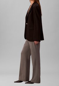 Femme portant un blazer noir à un bouton, un pantalon à jambes larges taupe et des chaussures noires pointues, debout de profil devant un fonds gris clair uni.