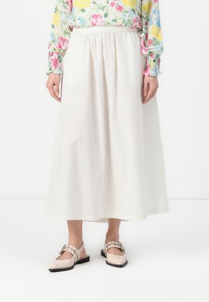 Kobieta ubrana w długą białą spódnicę, kwiecistą bluzkę z różowymi, żółtymi i zielonymi kwiatami oraz jasnoróżowe płaskie buty z klamrami.