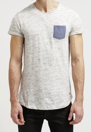 Uomo che indossa una t-shirt bianca a maniche corte con una tasca sul petto blu e pantaloni neri, in piedi contro uno sfondo chiaro e uniforme.