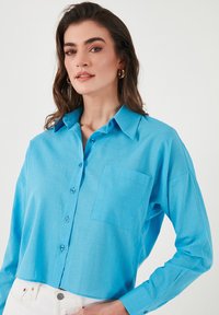 Light blue button-up shirt with a collar, long sleeves, a chest pocket, and a textured fabric, worn with white pants.