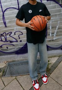 Camiseta negra de manga corta con los logos de Nike y Brooklyn, pantalones de mezclilla azul claro, sosteniendo un baloncesto naranja, zapatillas rojas y blancas.