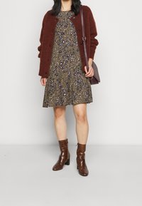 Ruskea, karvainen neuletakki kukkakuosisen, polvipituisen mekon päällä maanläheisissä sävyissä. Kiiltävät, ruskeat, pintaelävöitetyt nilkkasaappaat korkokengillä. Olkalaukku.