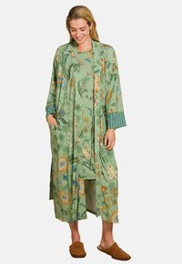 Vrouw in een groene bloempatronen ochtendjas en bijpassende nachtjapon met bruine sloffen, lachend en naar beneden kijkend.
