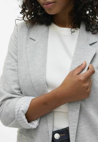 Hallikate blazer lõdva lõikega, millel on krae ja rullitud õlavarredega, valge pluusi kohal. Nähtav teksapantide vöökoht koos nööbiga.