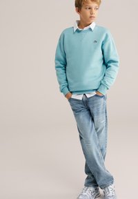 Chlapec s rukami v vreckách, oblečený v svetlomodrej mikine cez bielu košeľu, vyblednuté modré džínsy a sivé tenisky, stojaci na jednobarevnom pozadí.