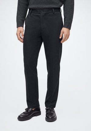 Homme portant un pantalon slim noir, des mocassins noirs à pampilles et un pull gris foncé, debout devant un fond uni.