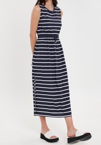 Robe maxi sans manches rayée bleu marine et blanche avec une taille élastique, des fentes sur les côtés, et associée à des sandales compensées blanches.